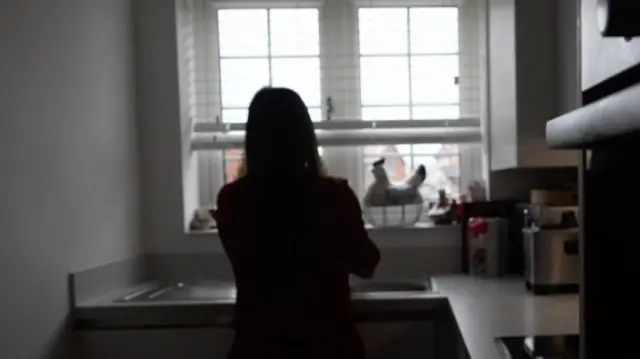Silhouette of a young woman standing by the kitchen sink with her back to us. 