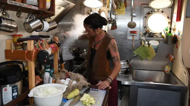 Giulia Baccosi na cozinha do navio, com seu cabelo escuro preso e avental marrom. Ela usa um colar e seu braço esquerdo tem várias tatuagens.