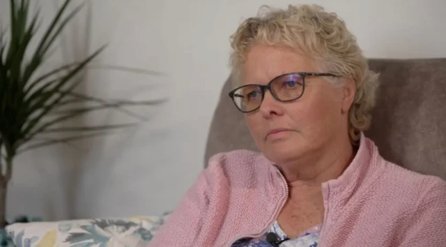 Gözlüklü ve sarı saçlı bir kadın kendisine soru soran gazeteciye bakıyor ve üzgün görünüyor.