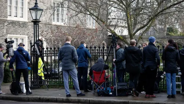 灰色の石造りの壁に白い木の窓枠の家を臨み、その前に黒い鉄柵で囲まれた緑地の手前に、ダウンジャケットなど防寒具を着た報道陣が複数、立っている。三脚でカメラを設置したり、組み立て式の簡易椅子に座って待機している人もいる。望遠レンズのついたカメラを携えている人もいる
