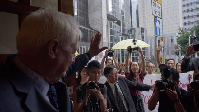 Hong Kong's former British colonial governor Chris Patten (L) waves at a small gathering of demonstrators (back, with umbrellas) as he arrives at the Foreign Correspondents' Club in Hong Kong on September 19, 2017.