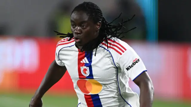 Tabitha Chawinga, visible jusqu'à la taille, est en pleine action. Elle porte un maillot blanc de Lyon orné d'une bande rouge et bleue au centre. Ses joues sont gonflées tandis qu'elle court pendant un match de football, ses nombreuses tresses flottant dans tous les sens derrière et sur les côtés de sa tête.