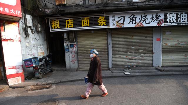A resident wearing a face mask and a plastic cap walks past closed shops in Wuhan, China