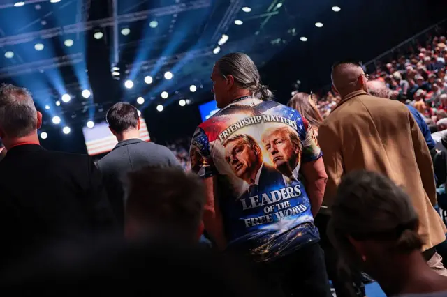 У великому залі з яскравим сценічним освітленням стоїть натовп людей. У центрі кадру — людина в футболці з великими портретами Орбана та Трампа з написом: "ЛІДЕРИ ВІЛЬНОГО СВІТУ". В тлі видно сцену, освітлювальні прожектори та заповнені трибуни. 