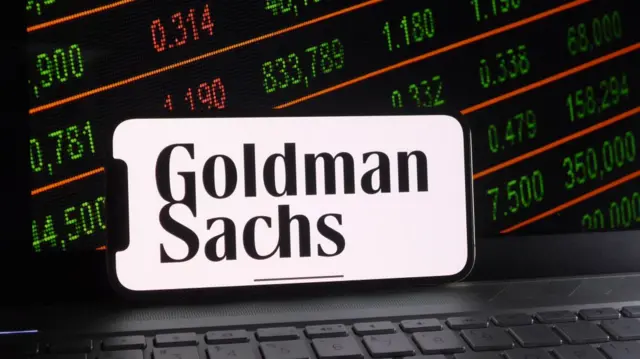 شعار غولدمان ساكس على هاتف ذكي وخلفه شاشة لسوق الأوراق المالية على كمبيوتر محمول