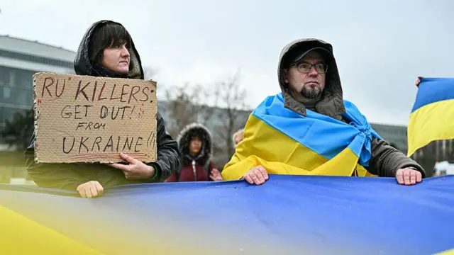 Протестувальники тримають плакати та українські прапори під час нового раунду переговорів між делегаціями Росії, України та США у Женеві, 17–18 лютого 2026 року.