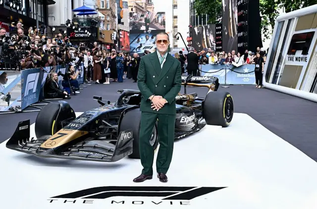 Brad Pitt yeşil bir takım elbiseyle bir F1 aracının önünde ellerini önünde bağlayıp poz veriyor
