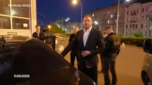 Знімок екрана відео: на передньому плані – чорне авто. Поруч стоїть Єрмак у темному костюмі та світлій сорочці. Праворуч видно людину в куртці з великим написом «НАБУ» на спині. У кутку кадру є напис «11.05.2026».
