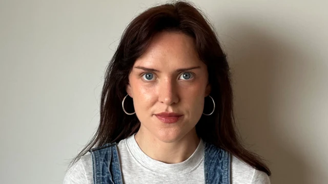 Keelin mira fijamente a la cámara. Tiene ojos azules y aros plateados, y lleva el pelo castaño suelto. Lleva una camiseta gris y se le ven los tirantes de su overol. Está de pie contra una pared blanca.