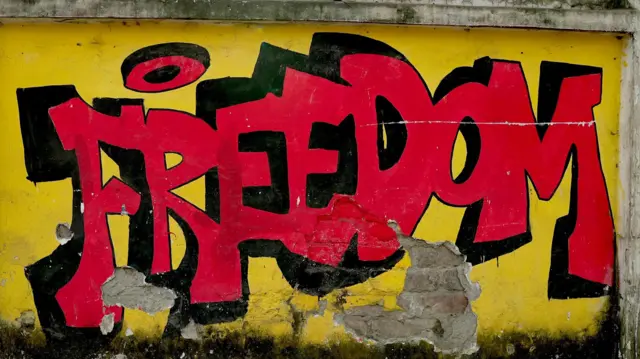 Um mural pintado traz a palavra “Liberdade”, em letras vermelhas sobre fundo amarelo 