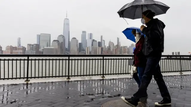 Duas pessoas usando máscaras caminham sob a chuva segurando guarda-chuvas com a cidadenova york ao fundo, do outro lado do rio