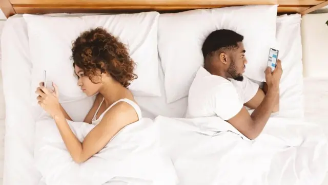 رجل وامرأة متجافيين على سرير واحد وكلٌّ يتصفح هاتفه