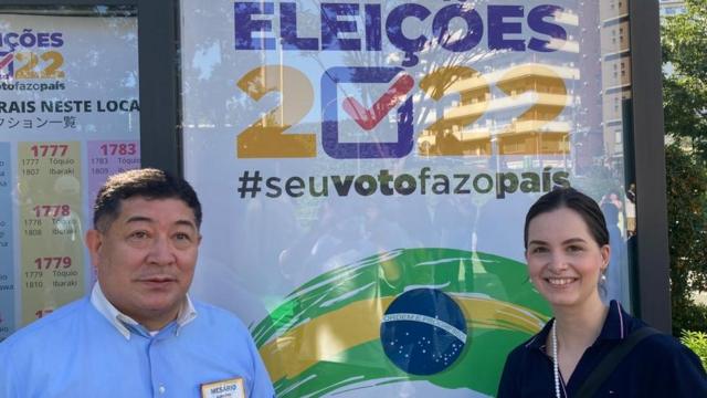 Fotografia colorida mostra um homemgrupo de sinais pixbetaparência nipônica ao ladogrupo de sinais pixbetuma mulher brancagrupo de sinais pixbetfrente a um poster escrito eleições 2022