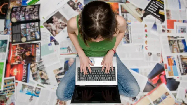 Mulher sentada no chão usando laptop rodeada por revistas e jornais