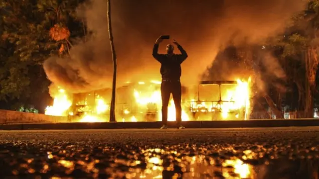 Un hombre se toma una selfie frente a un autobús en llamas.