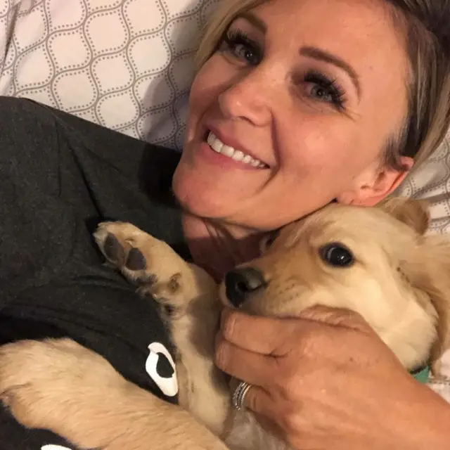 Paula deitada na cama segurando o cachorrinho Todd. Ela está sorrindo. Ele parece estar relaxado.