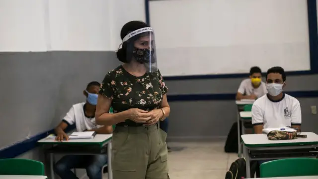 Professoracbet levelspé com máscara e protetor facial. Alunos, que estão sentados, usam mácaras e uniforme.