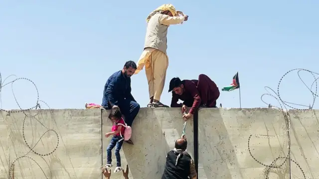 Afghans try to enter Kabul airport, with a men pulling a child up the side of a wall while another man pulls a man nearby