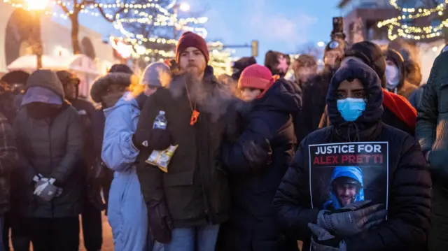 Множество людей, на переднем плане смуглый молодой человек в капюшоне и медицинской маске, он держит плакат с надписью Justice for Alex Pretti.