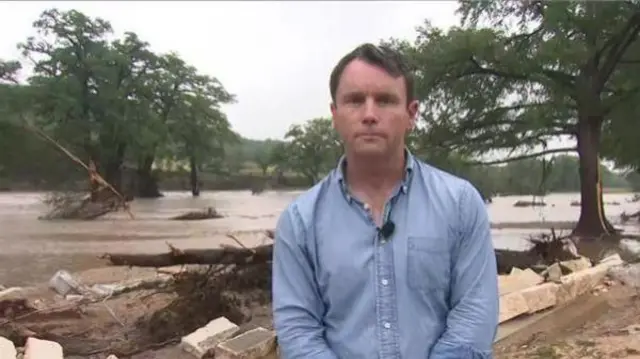 水位が上昇した川を背に、水色のシャツを着た男性記者がカメラに向かって立っている。後ろには、なぎ倒された樹木や、崩れたブロックなどが散乱している