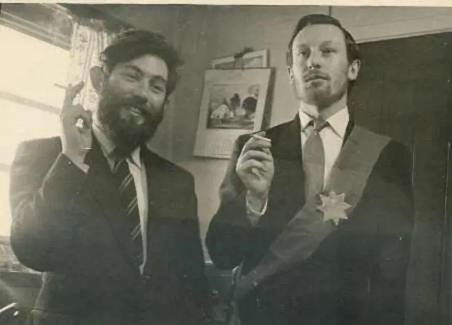 Dois homens vestindo ternos e gravatas e sorrindo para a câmera