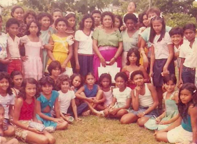 Dona Onete posa para foto cercada por crianças; ela veste uma camisa verde e uma saia rosa, e segura uma sacola.