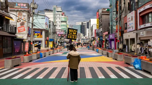 Uma rua de pedestres animada e colorida em Seul, repleta de faixas de pedestres vibrantes, lojas exclusivas e muita atividade. Uma mulher está no meio de uma faixa de pedestres, vestindo uma jaqueta de inverno e carregando a placa "Sala de Escape, metade do preço".