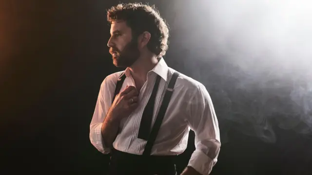 Ben Platt se encuentra bajo una iluminación teatral dramática. Lleva una elegante camisa blanca con el cuello abierto, tirantes y una corbata negra desatada alrededor del cuello. Mira a un lado, a lo lejos. Tiene cabello y barba oscuros, y una luz brilla sobre su hombro izquierdo.