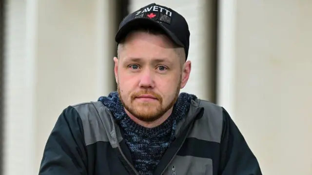 Homem na casa dos 30 anos de idade olha de frente para a câmera. Ele veste um casaco e usa um boné de baseball com o nome 'Zavetti'.