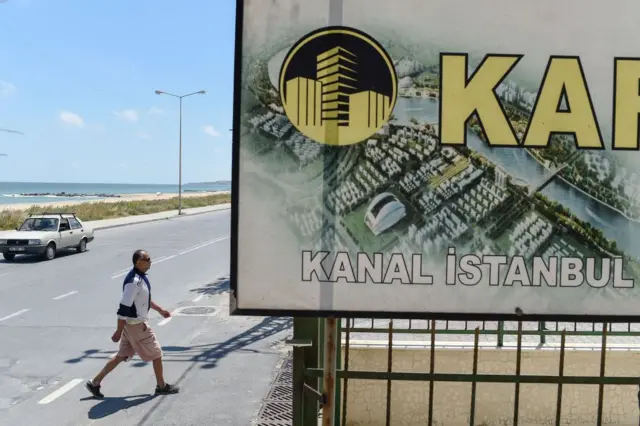 Kanal İstanbul çevresine yapılacak inşaatların çizimini gösteren bir pano, yanında bir erkek yürüyor, arkada deniz gözüküyor