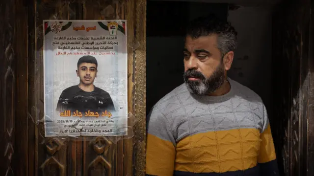 يقف والد جاد بجوار ملصق تذكاري لابنه، مُلصق على الباب الأمامي الخشبي المنحوت للعائلة في مخيم الفارعة للاجئين، في الضفة الغربية المحتلة