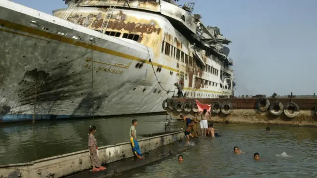 বন্দরে শিশুরা সাঁতার কাটছে, আর অন্যরা একটি বিশাল, মরিচা ধরা জাহাজের পাশে  দাঁড়িয়ে আছে। যার সামনের অংশে সোনালী অক্ষরে 'আল মনসুর' লেখা। বন্দরের জেটি থেকে টায়ার ঝুলছে।