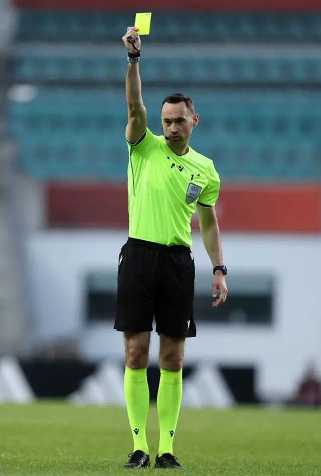 El árbitro Tom Owens, con camisa y medias verdes y pantaloneta negra, estira el brazo hacia arriba con una tarjeta amarilla en Estonia durante un partido el 4 de junio de 2026

