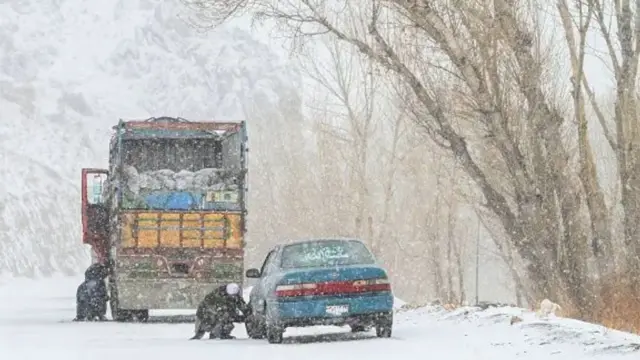 تصویری از یک موتر نوع کرولا و یک موتر لاری که کنار جاده ایستاده‌اند و در حال بستن زنجیبر به تایرهای موتر برای عبور در مسیر برفی هستند.