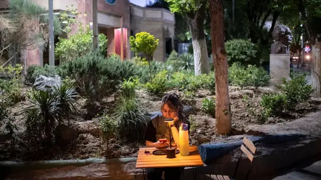 یک زن جوان ایرانی در حالی که در یک کافه روباز در مرکز شهر تهران، ایران نشسته است، در ۱۱ آوریل ۲۰۲۶ (۲۲ فروردین ۱۴۰۵) از تلفن همراه خود استفاده می‌کند، در حالی که مذاکرات مستقیم ایران و ایالات متحده در اسلام‌آباد، پاکستان در جریان است.