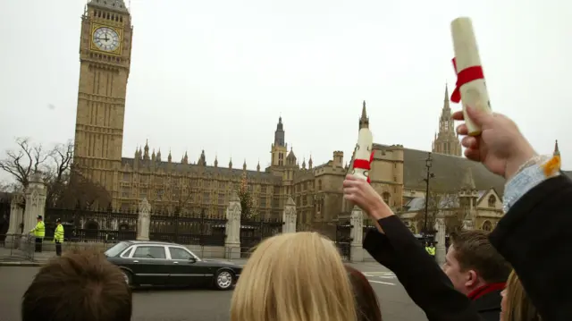 Wide view of a university, people standing beneath what looks like the building and showing their wrapped certificate