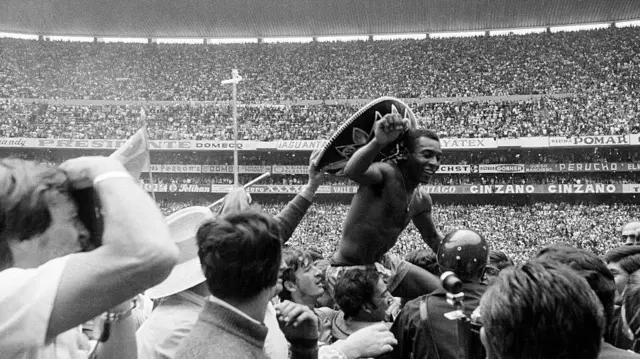 A foto em preto e branco mostra Pelé sendo carregado nos ombros por uma multidão dentro de um estádio lotado. Sem camisa e usando um grande sombrero, ele ergue o dedo indicador enquanto sorri para o público, cercado por torcedores, fotógrafos e seguranças. Ao fundo, as arquibancadas completamente cheias e os painéis de publicidade reforçam o clima de celebração após uma partida.
