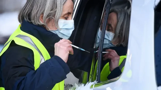 یک کارمند بهداشتی در فضای باز در حال تزریق واکسن به شخصی که در خودروی خود نشسته است