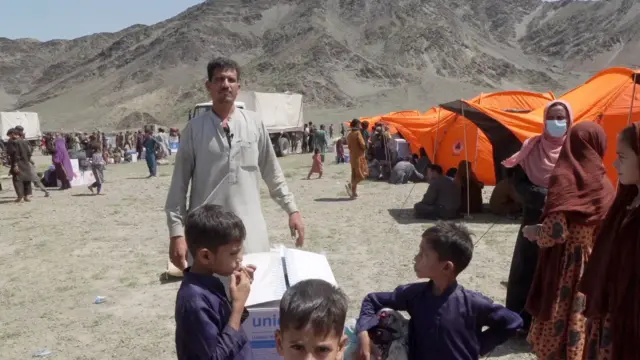 مردی با موهای کوتاه تیره و سبیل در فضای باز با پیراهن به رنگ بژ ایستاده است. کودکانی با پیراهن‌های بنفش در مقابل او، چادرهای نارنجی در سمت راست و کوه‌های خاکستری در پس زمینه دیده می‌شوند