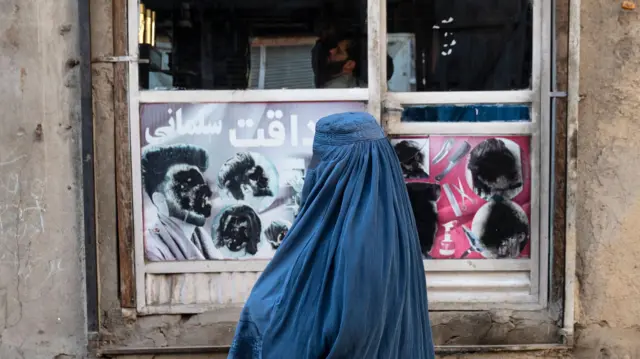 در تصویر یک زن افغان با برقع از کنار یک آرایشگاه مردانه با تصاویر مخدوششده مردان در شهر کابل عبور میکند
