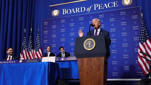 US President Donald Trump, in a navy suit and a purple tie, speaks at the inaugural Board of Peace meeting. The background reads "Board of Peace"