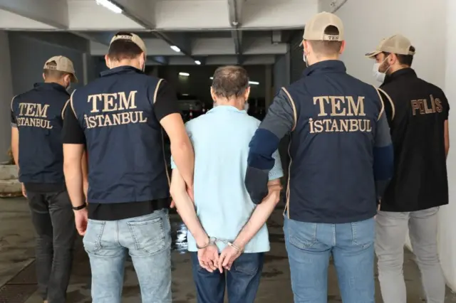 Mavi tişörtlü bir kişi, elleri ters kelepçeli şekilde polis tarafından götürülüyor. 