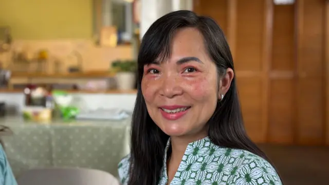 喬伊斯·威爾莫特（Joyce Wilmot）望向鏡頭微笑，她擁有長黑的頭髮，並穿上綠白相間的恤衫。