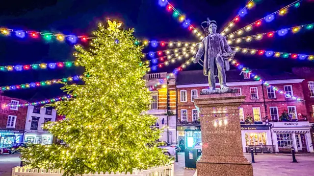 Un árbol de Navidad iluminado con luces blancas. Está en una plaza rodeada de edificios de ladrillo rojo. Hay una estatua de un hombre sobre un pedestal y luces multicolores en lo alto.