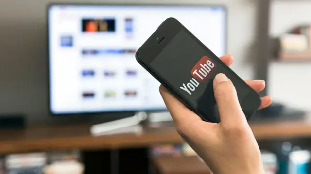 Una mano sostiene un móvil con la aplicación de YouTube frente a una pantalla de televisión