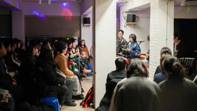 一個房間裡，幾個年輕人站在話筒後面，面向很多坐在椅子上的人。