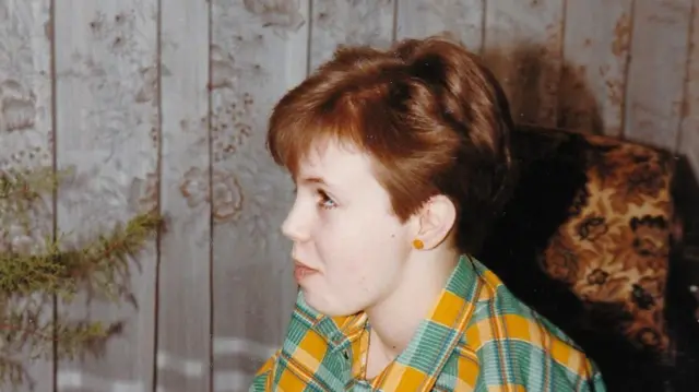 Egy 1982-ben készült színes fényképen a 11 éves Nina látható, amint egy nappaliban kerekesszékben ül. Zöld-sárga kockás összeillő ruhát visel egy világos színű, hosszú ujjú felső fölött, és egy narancssárga gyöngysort tart a kezében. A háttérben egy ablakot csipkefüggöny takar, a kerekesszék mellett pedig részben egy mintás fotel látható.