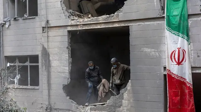 ミサイル攻撃で激しく損傷した住宅の壁や窓がなくなった部分で、マスクを着けた2人が立っている。近くにイラン国旗が見える