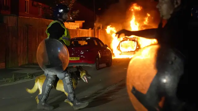 Policial passa com cãobabado betanorua com um carrobabado betanochamas ao fundo 