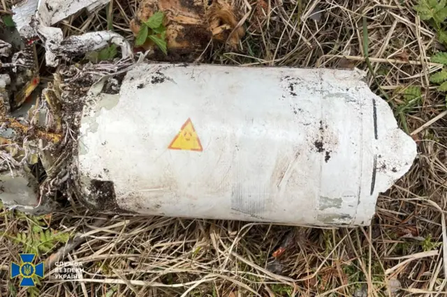 Пошкоджений металевий циліндричний предмет, що лежить на землі серед сухої трави й уламків. На корпусі видно жовтий попереджувальний знак у вигляді трикутника, а з одного краю – обірвані дроти й внутрішні деталі.